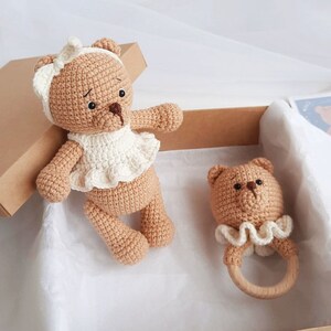 Puede incluir: Dos osos de peluche tejidos a crochet, uno con un vestido blanco y el otro con un sonajero. Los osos son marrones y tienen ojos negros. El sonajero es de madera y tiene un borde blanco tejido a crochet.
