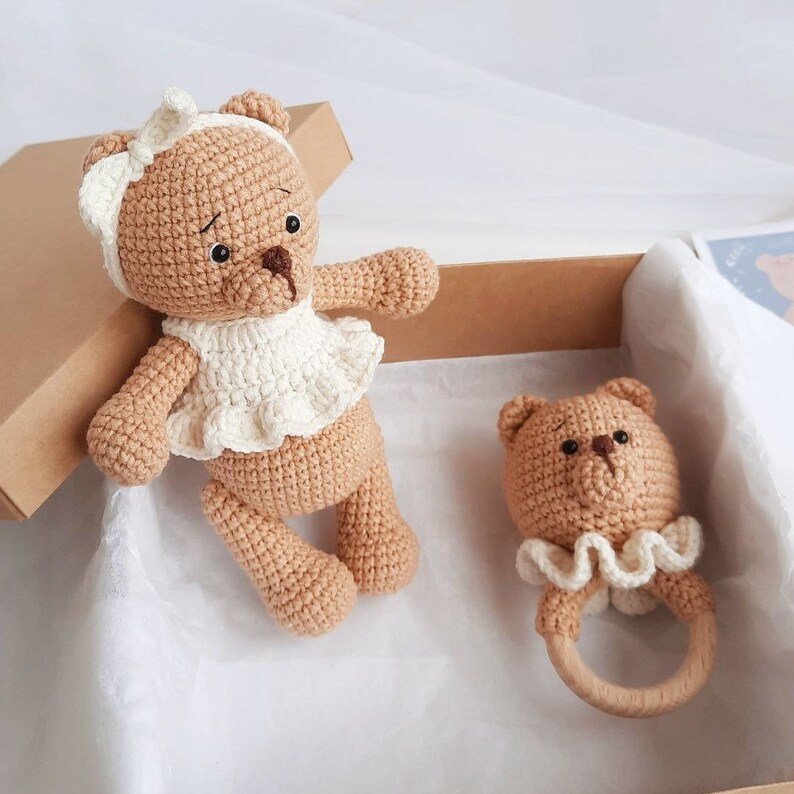 Puede incluir: Dos osos de peluche tejidos a crochet, uno con un vestido blanco y el otro con un sonajero. Los osos son marrones y tienen ojos negros. El sonajero es de madera y tiene un borde blanco tejido a crochet.