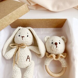 May include: A cream-coloured crochet bunny and bear baby toy set in a gift box. The bunny has long ears, a bow, and the letter "A" embroidered on its body. The bear has a wooden ring and a ruffled collar.