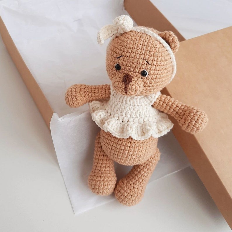 Puede incluir: Un oso de peluche de crochet marr&oacute;n que lleva un vestido blanco de crochet con un lazo blanco en la cabeza. El oso est&aacute; sentado en una caja marr&oacute;n.