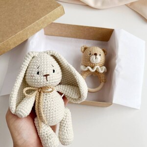 May include: A crochet bunny and bear rattle set in a kraft gift box. The bunny is off-white with long ears and a tan bow. The bear rattle is tan with a wooden ring and a cream ruffle. A perfect baby gift.
