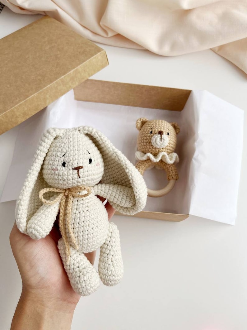 May include: A crochet bunny and bear rattle set in a kraft gift box. The bunny is off-white with long ears and a tan bow. The bear rattle is tan with a wooden ring and a cream ruffle. A perfect baby gift.