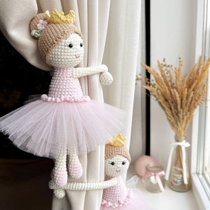 Op de afbeelding: Twee gehaakte ballerinapoppen met roze tutu's en gouden kronen worden gebruikt als gordijnbandjes. De poppen houden een wit gordijn vast.