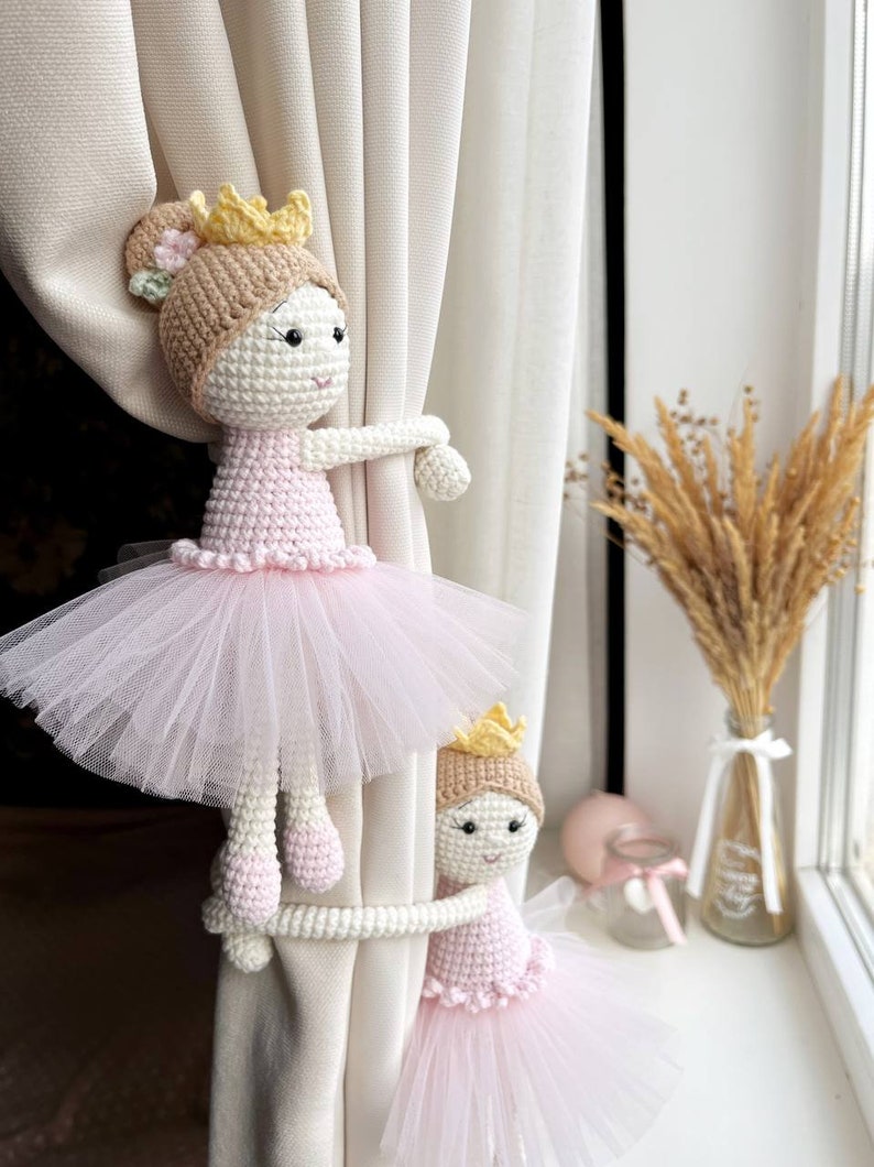 Op de afbeelding: Twee gehaakte ballerinapoppen met roze tutu's en gouden kronen worden gebruikt als gordijnbandjes. De poppen houden een wit gordijn vast.