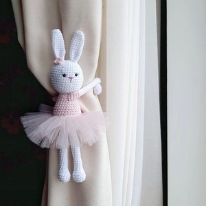 May include: A white crocheted bunny wearing a pink tutu and holding a curtain tieback. The bunny is holding the tieback with its right arm and has a pink flower on its head.