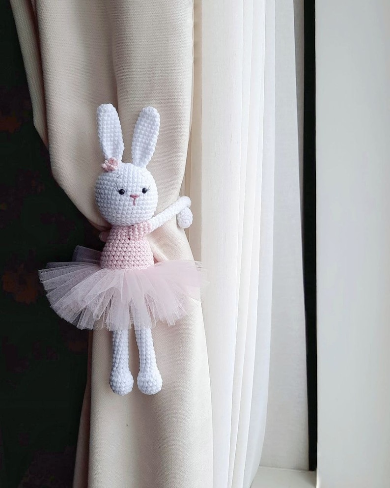 May include: A white crocheted bunny wearing a pink tutu and holding a curtain tieback. The bunny is holding the tieback with its right arm and has a pink flower on its head.