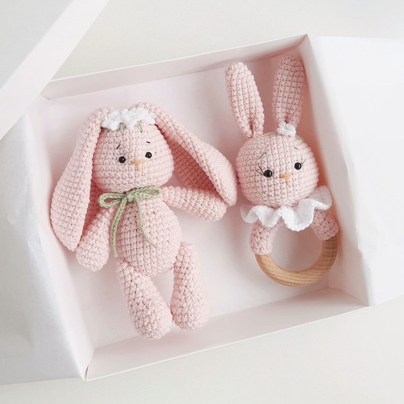 May include: Two pink crocheted bunny toys in a white gift box. One bunny is a rattle with a wooden ring and a white crocheted collar. The other bunny has a green ribbon around its neck.