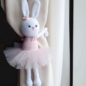 Puede incluir: Un conejito de crochet blanco con un tut&uacute; rosa y agarrado a una cortina. El conejito tiene ojos negros y una nariz rosa.