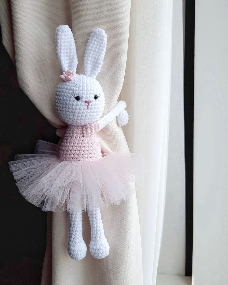 Puede incluir: Un conejito de crochet blanco con un tut&uacute; rosa y agarrado a una cortina. El conejito tiene ojos negros y una nariz rosa.