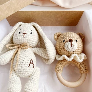 May include: A cream-coloured crocheted bunny with long ears and a brown letter "A" on its chest, and a tan bear rattle with a wooden ring. The set is presented in a brown gift box, ideal for a baby.