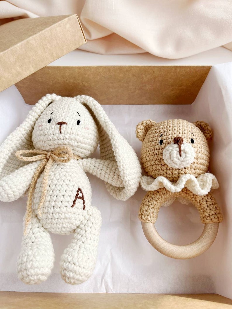 May include: A cream-coloured crocheted bunny with long ears and a brown letter "A" on its chest, and a tan bear rattle with a wooden ring. The set is presented in a brown gift box, ideal for a baby.
