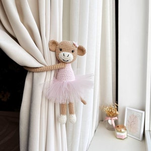 May include: A crocheted monkey doll wearing a pink tutu, holding onto a white curtain. The monkey has brown fur, a pink bow, and white shoes.