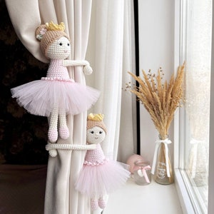 Op de afbeelding: Twee gehaakte ballerinapoppen met roze tutu's en gouden kronen worden gebruikt als gordijnbandjes op een wit gordijn.