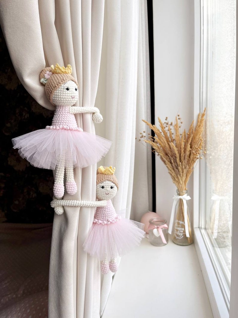 Op de afbeelding: Twee gehaakte ballerinapoppen met roze tutu's en gouden kronen worden gebruikt als gordijnbandjes op een wit gordijn.