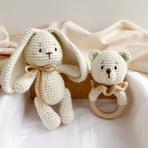 May include: A cream-coloured crocheted bunny and bear rattle set. The bunny has long ears and a bow, while the bear has a wooden ring. Both are handmade, ideal as a baby gift.