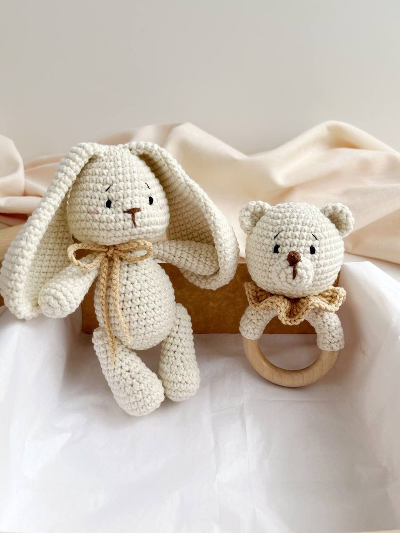 May include: A cream-coloured crocheted bunny and bear rattle set. The bunny has long ears and a bow, while the bear has a wooden ring. Both are handmade, ideal as a baby gift.