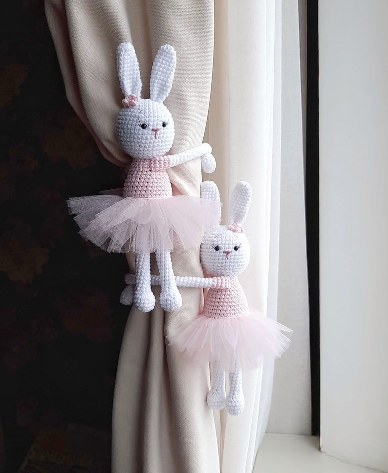 Puede incluir: Dos mu&ntilde;ecas de conejo de crochet blanco vestidas con tut&uacute;s rosas se utilizan como ataduras de cortinas. Los conejos sostienen las cortinas beige con sus brazos.