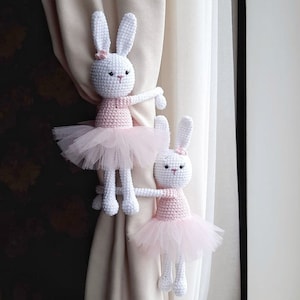 May include: Two white crocheted bunny curtain tiebacks with pink tutus. The bunnies are holding onto the curtain with their arms.