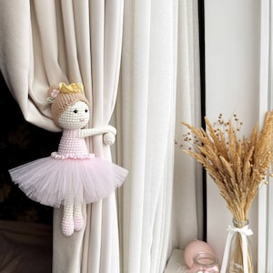 Op de afbeelding: Een gehaakte ballerinapop met een roze tutu en een gouden kroon, die aan een witte gordijn vasthoudt. De pop is een gordijnband.