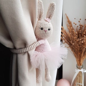 May include: A crocheted bunny doll wearing a pink tutu, used as a curtain tieback. The bunny has black eyes and a pink nose.