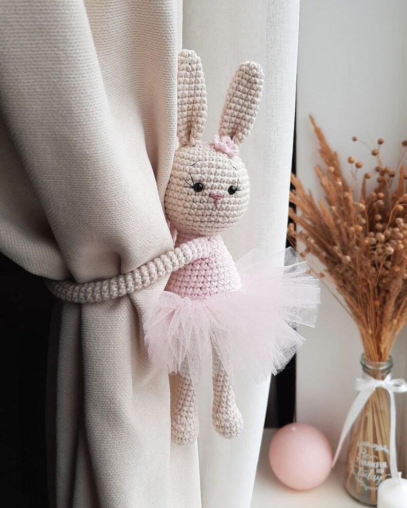 May include: A crocheted bunny doll wearing a pink tutu, used as a curtain tieback. The bunny has black eyes and a pink nose.