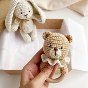 May include: Crocheted baby toys. A beige bunny with long ears and a tan bear rattle with a wooden ring. The bear has a white ruffled collar. The toys are in a cardboard box.