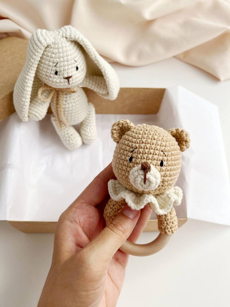 May include: Crocheted baby toys. A beige bunny with long ears and a tan bear rattle with a wooden ring. The bear has a white ruffled collar. The toys are in a cardboard box.
