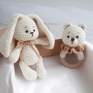 May include: A white crocheted bunny and a white crocheted bear with a wooden ring rattle. Both toys have brown eyes and noses.