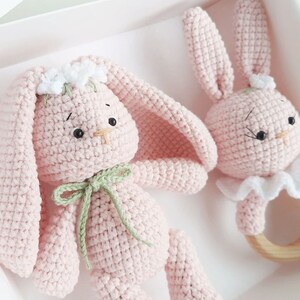 May include: Two pink crocheted bunny toys. One bunny is a rattle with a wooden ring and a white ruffle. The other bunny has a green ribbon around its neck and white flowers on its head.