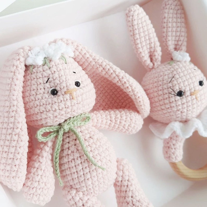 May include: Two pink crocheted bunny toys. One bunny is a rattle with a wooden ring and a white ruffle. The other bunny has a green ribbon around its neck and white flowers on its head.