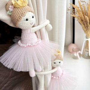 Op de afbeelding: Twee gehaakte ballerinapoppen met roze tutu's en gouden kronen worden gebruikt als gordijnbandjes. De poppen houden een witte gordijn vast.