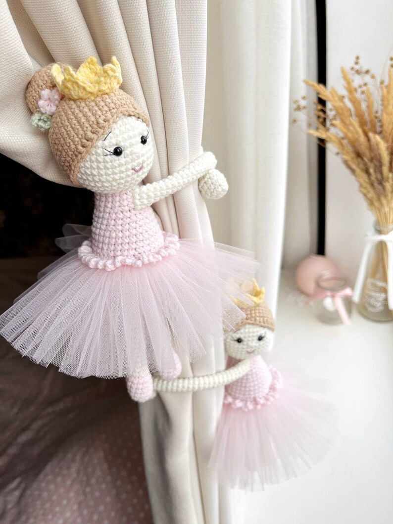 Op de afbeelding: Twee gehaakte ballerinapoppen met roze tutu's en gouden kronen worden gebruikt als gordijnbandjes. De poppen houden een witte gordijn vast.
