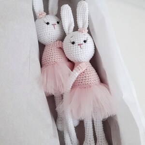 Puede incluir: Dos mu&ntilde;ecas de conejo de crochet blanco con tut&uacute;s rosas. Los conejos tienen ojos negros y narices rosas. Est&aacute;n sentados en una caja de regalo blanca.