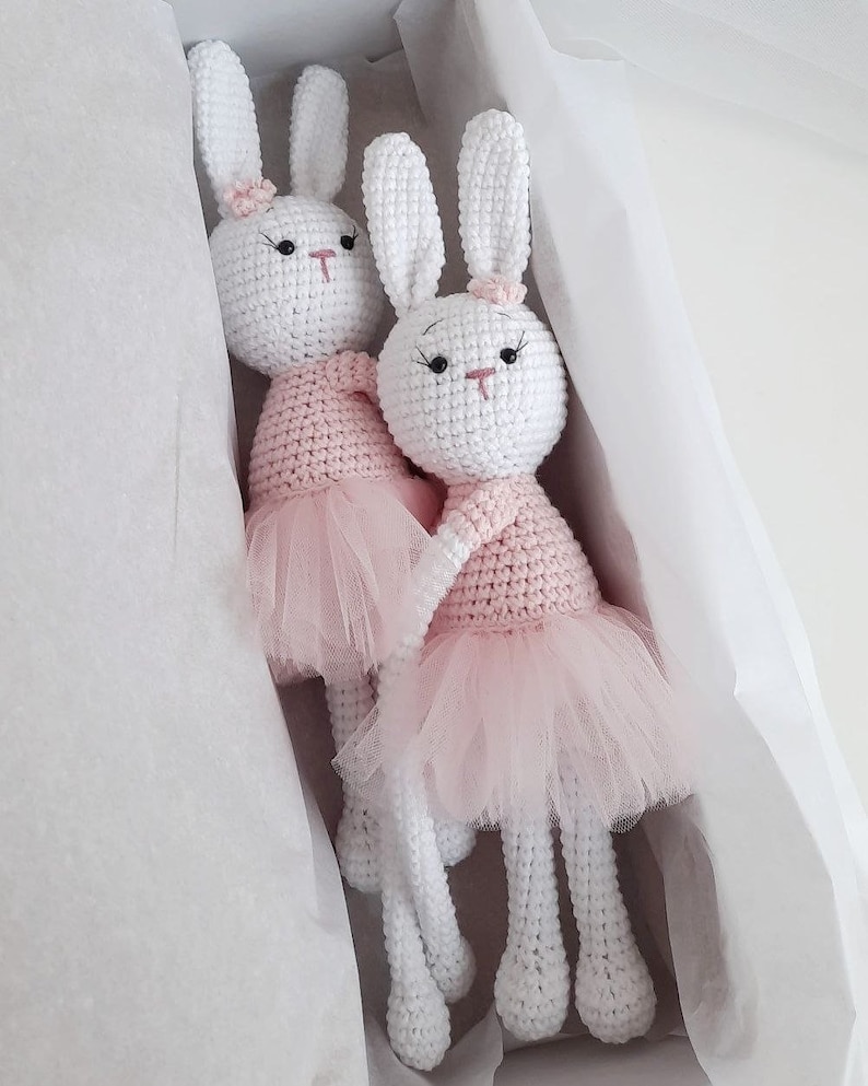 Puede incluir: Dos mu&ntilde;ecas de conejo de crochet blanco con tut&uacute;s rosas. Los conejos tienen ojos negros y narices rosas. Est&aacute;n sentados en una caja de regalo blanca.