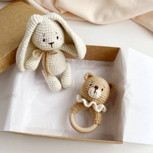 May include: A crochet bunny and bear baby rattle set in a cardboard box. The bunny is off-white with long ears and a tan bow. The bear rattle is tan with a white frilly collar and a wooden ring.