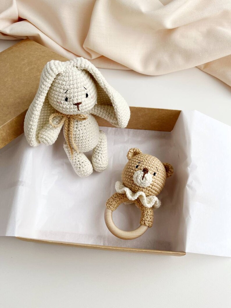 May include: A crochet bunny and bear baby rattle set in a cardboard box. The bunny is off-white with long ears and a tan bow. The bear rattle is tan with a white frilly collar and a wooden ring.