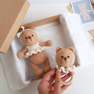 Puede incluir: Dos osos de peluche de crochet marrones, uno con un vestido de crochet blanco y el otro con un babero de crochet blanco y un anillo de dentici&oacute;n de madera. Los osos est&aacute;n en una caja marr&oacute;n con papel de seda blanco.