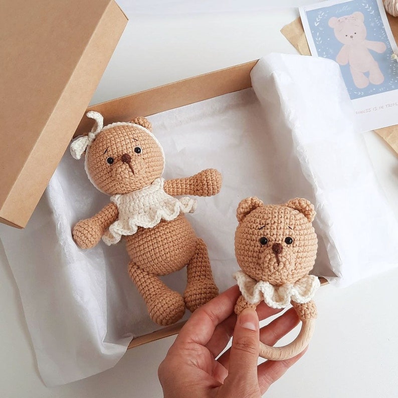 Puede incluir: Dos osos de peluche de crochet marrones, uno con un vestido de crochet blanco y el otro con un babero de crochet blanco y un anillo de dentici&oacute;n de madera. Los osos est&aacute;n en una caja marr&oacute;n con papel de seda blanco.