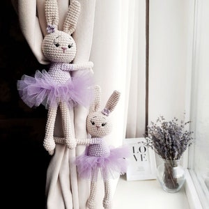 May include: Two crocheted bunny rabbit curtain tiebacks with purple tutus. The bunnies are white with black eyes and pink noses. The tutus are made of sheer purple tulle.