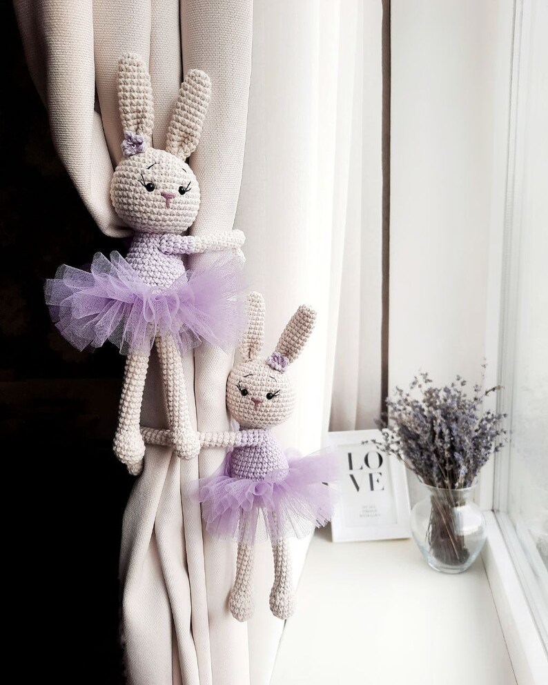 May include: Two crocheted bunny rabbit curtain tiebacks with purple tutus. The bunnies are white with black eyes and pink noses. The tutus are made of sheer purple tulle.
