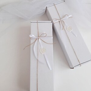 Puede incluir: Dos cajas de regalo blancas atadas con cuerda y cinta blanca. Cada caja tiene un sello de cera blanco con un dise&ntilde;o.