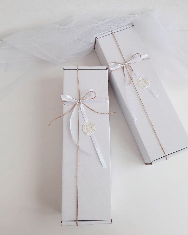 Puede incluir: Dos cajas de regalo blancas atadas con cuerda y cinta blanca. Cada caja tiene un sello de cera blanco con un dise&ntilde;o.