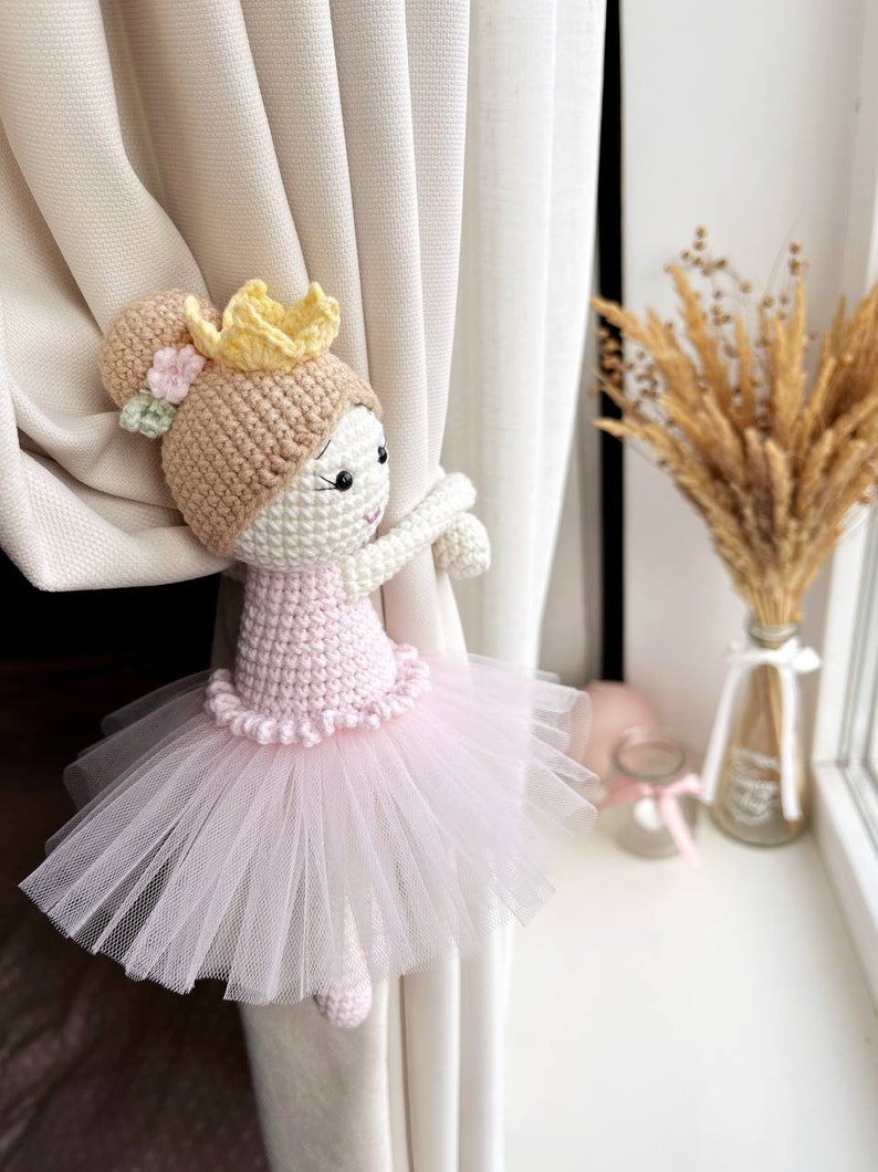 Op de afbeelding: Een gehaakte ballerinapop met een roze tutu en een gele kroon, die aan een wit gordijn vasthoudt.
