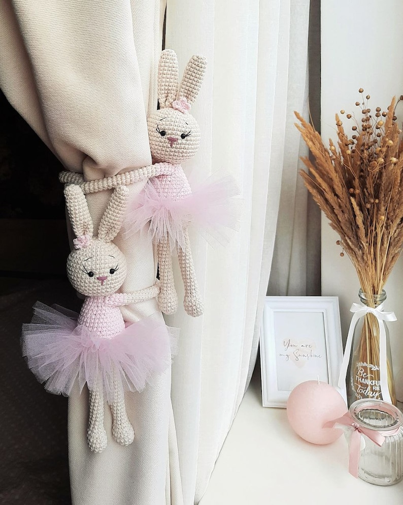 Puede incluir: Dos mu&ntilde;ecas de conejo de crochet vestidas con tut&uacute;s rosas se utilizan como ataduras de cortinas. Los conejos se agarran a una cortina blanca con sus brazos. Los conejos est&aacute;n sobre un fondo blanco.