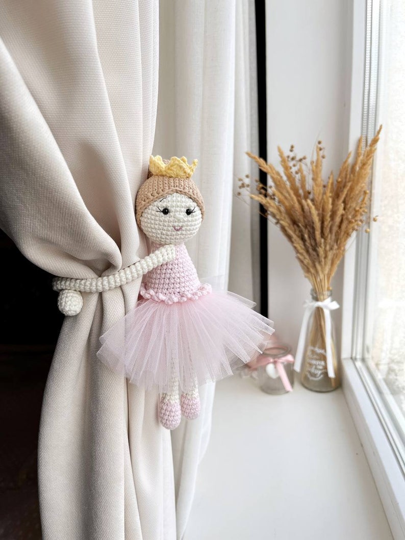 Op de afbeelding: Een gehaakte ballerinapop met een roze tutu en een gele kroon, gebruikt als gordijnband.
