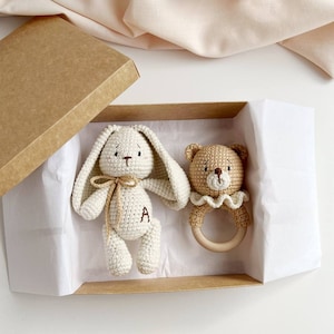 May include: A crochet baby gift set in a kraft box. The set includes a cream-coloured bunny with a bow and the letter "A" on its leg, and a tan bear rattle with a wooden ring. The box is lined with white paper.