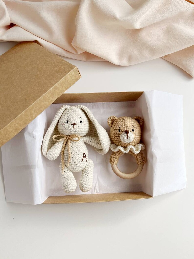 May include: A crochet baby gift set in a kraft box. The set includes a cream-coloured bunny with a bow and the letter "A" on its leg, and a tan bear rattle with a wooden ring. The box is lined with white paper.