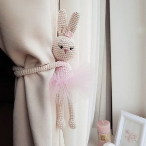 May include: A crocheted bunny wearing a pink tutu, used as a curtain tieback. The bunny is holding onto a white curtain with its paws.