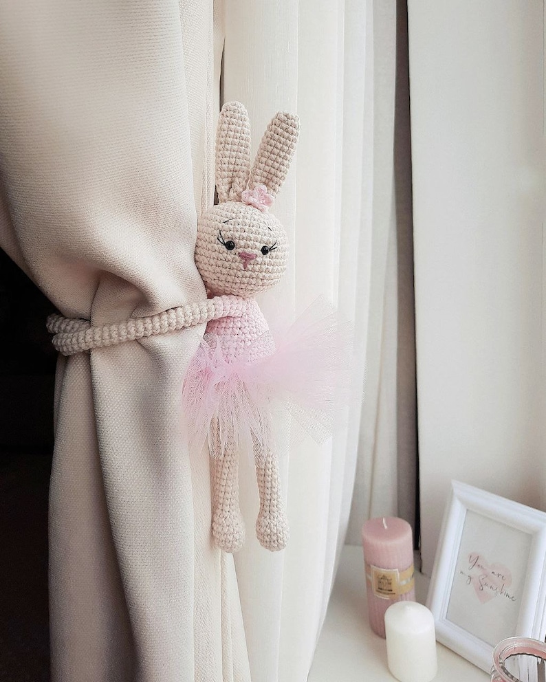May include: A crocheted bunny wearing a pink tutu, used as a curtain tieback. The bunny is holding onto a white curtain with its paws.