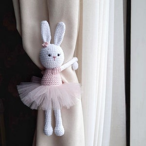 Puede incluir: Un conejito de crochet blanco con un tut&uacute; rosa y sujetando un amarrador de cortina. El conejito est&aacute; sujetando el amarrador con su brazo derecho y tiene una flor rosa en la cabeza.